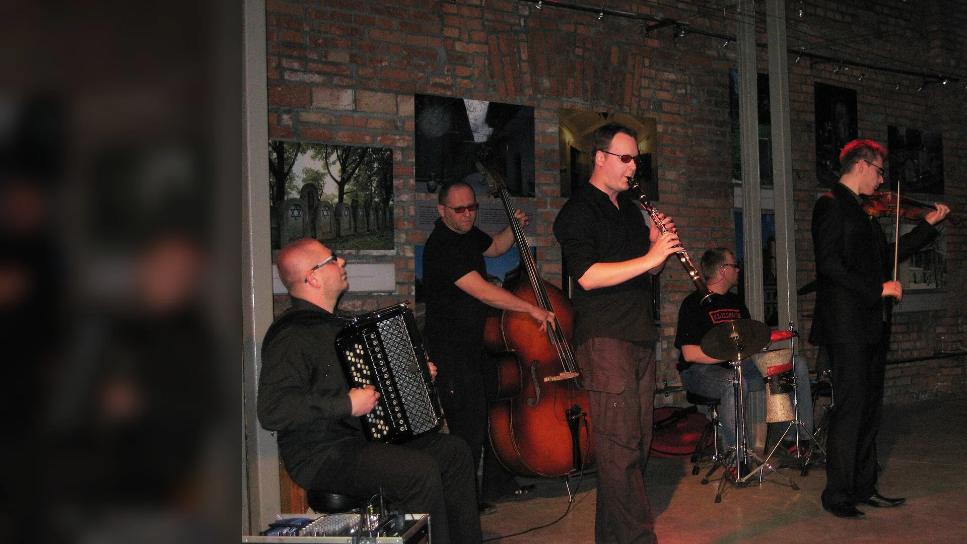 A klezmer quintet plays accordion, double bass, clarinet, drums and violin on a small stage in front of a brick wall hung with photographs.