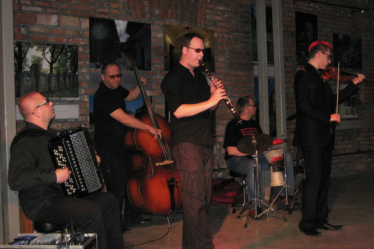 A klezmer quintet plays accordion, double bass, clarinet, drums and violin in front of a brick wall hung with photographs.