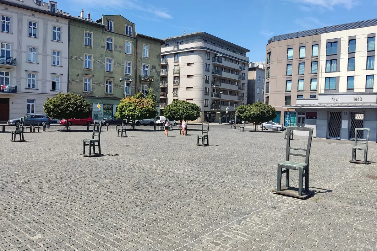 Large empty chairs stand on a cobbled square lined with apartment buildings under a blue sky.